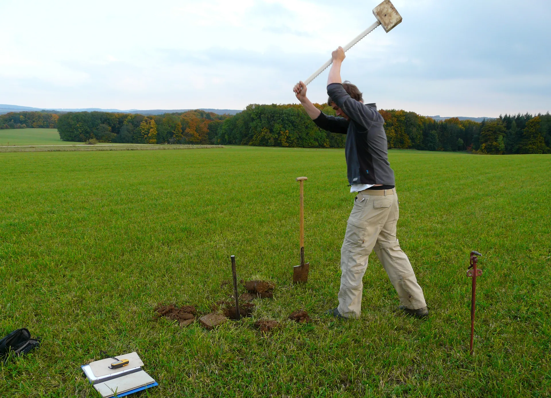 Auf einer Wiese schlägt ein Mann einen Bohrstock mit einem großen Hammer in den Boden. Der Oberboden wurde zuvor spatentief ausgehoben.