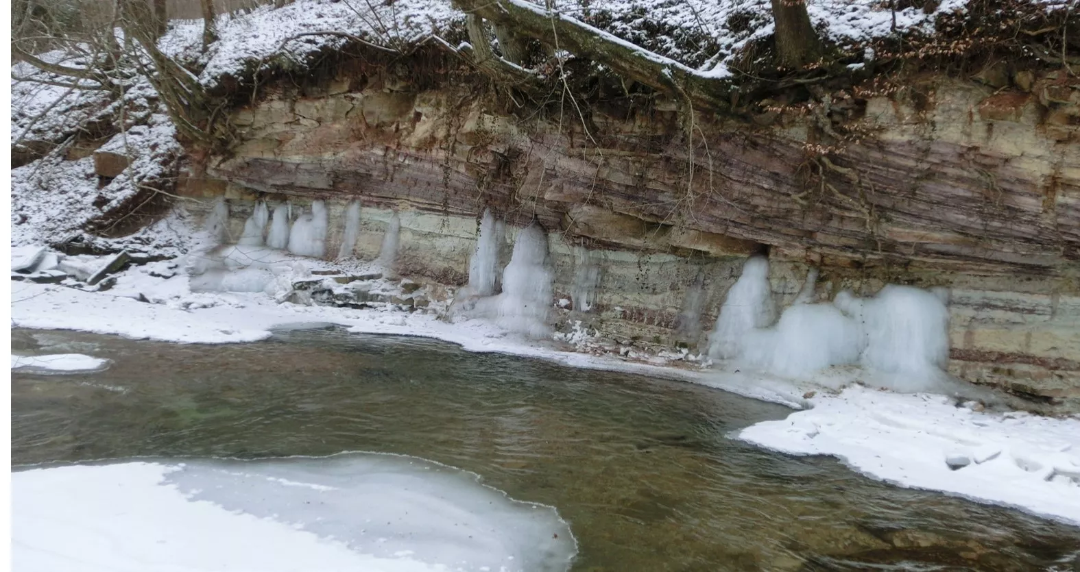 Fluss in einer winterlich eingeschneiten Umgebung. Das Ufer bildet eine Felswand. Aus einer waagerechten Spalte dringt zu Eis gefrorenes Wasser.
