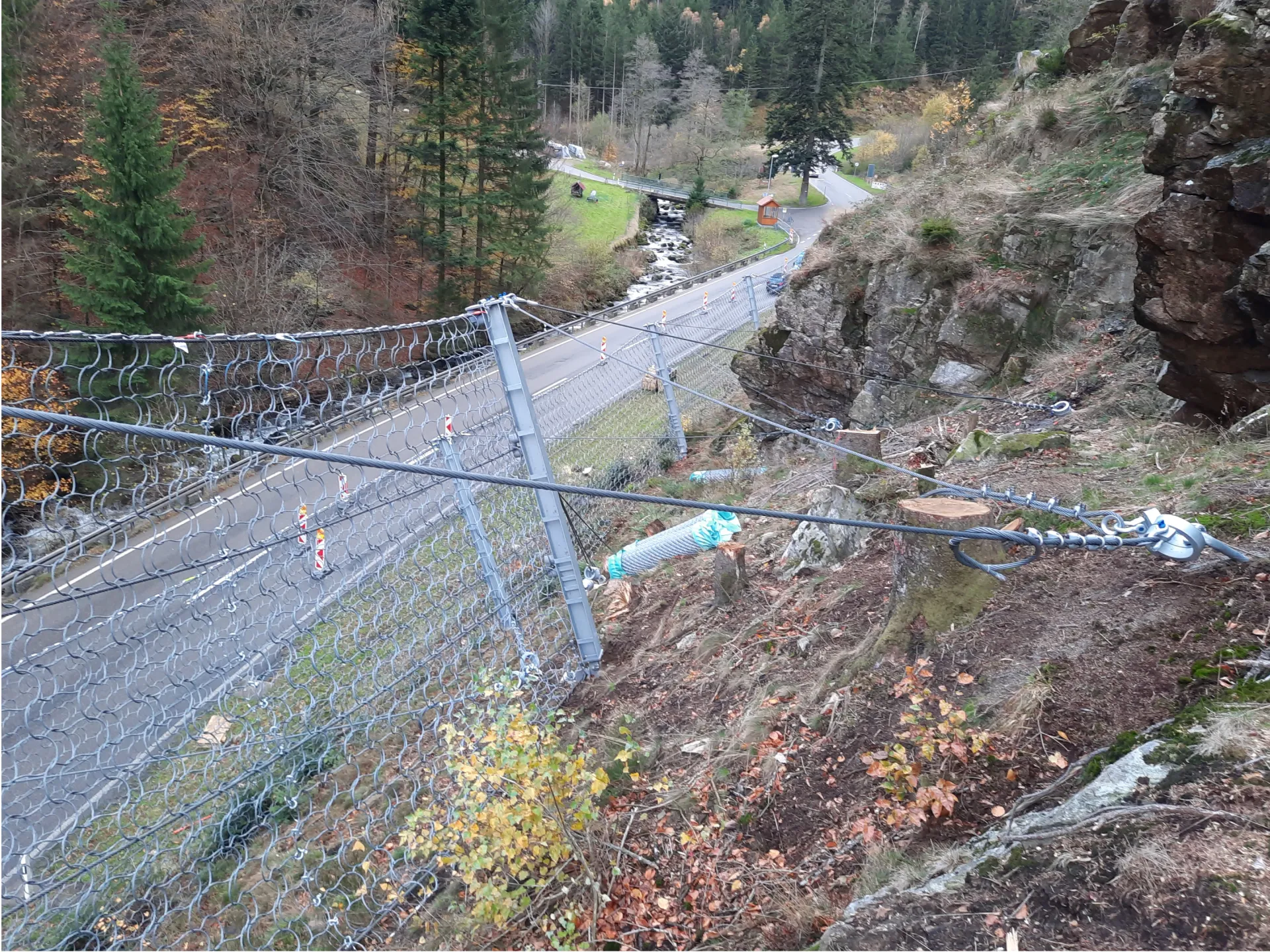 Talwärtiger Blick auf einen Steinschlagschutzzaun mit zahlreichen Stützen, Drahtseilen und Zaungeflecht. Im Hintergrund des im Hangelände stehenden Zauns ist die darunterliegende Landestraße sichtbar.