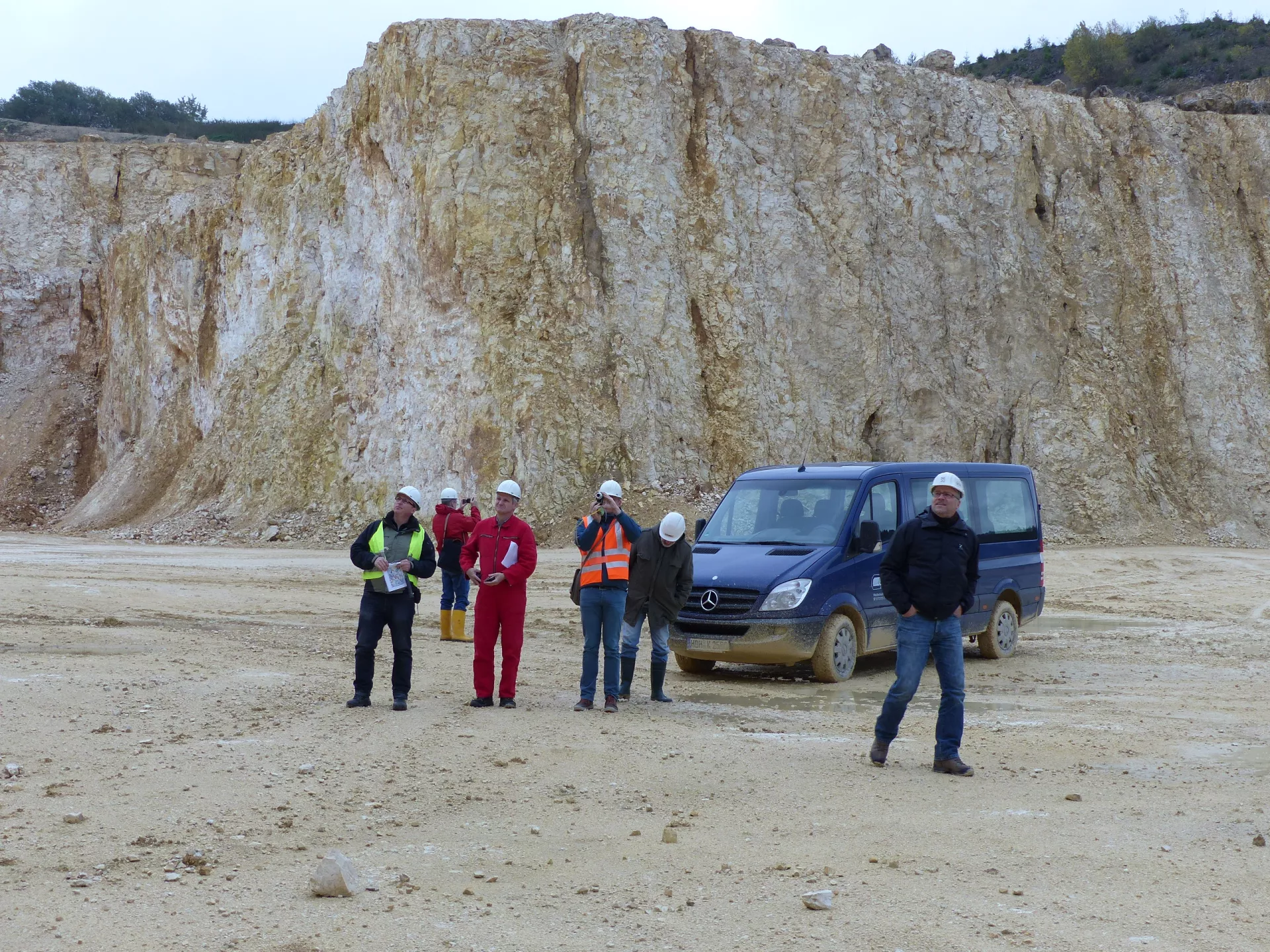 Mehrere Geologen stehen in einem Steinbruch. Im Hintergrund ist eine mehrere zehner Meter hohe Felswand.
