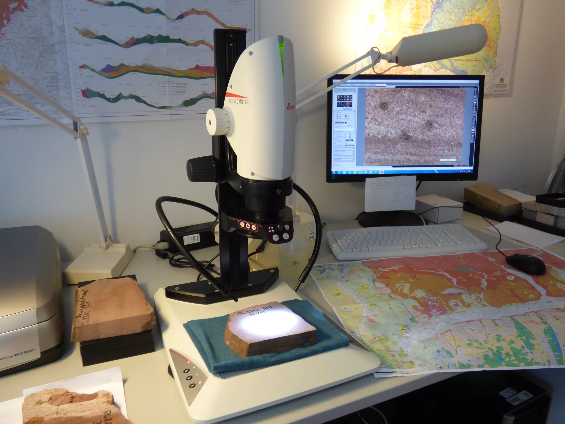 Eine rötliche Sandsteinprobe liegt unter einem Fotomikroskop und wird vergrößert an einem PC-Bildschirm angezeigt. 