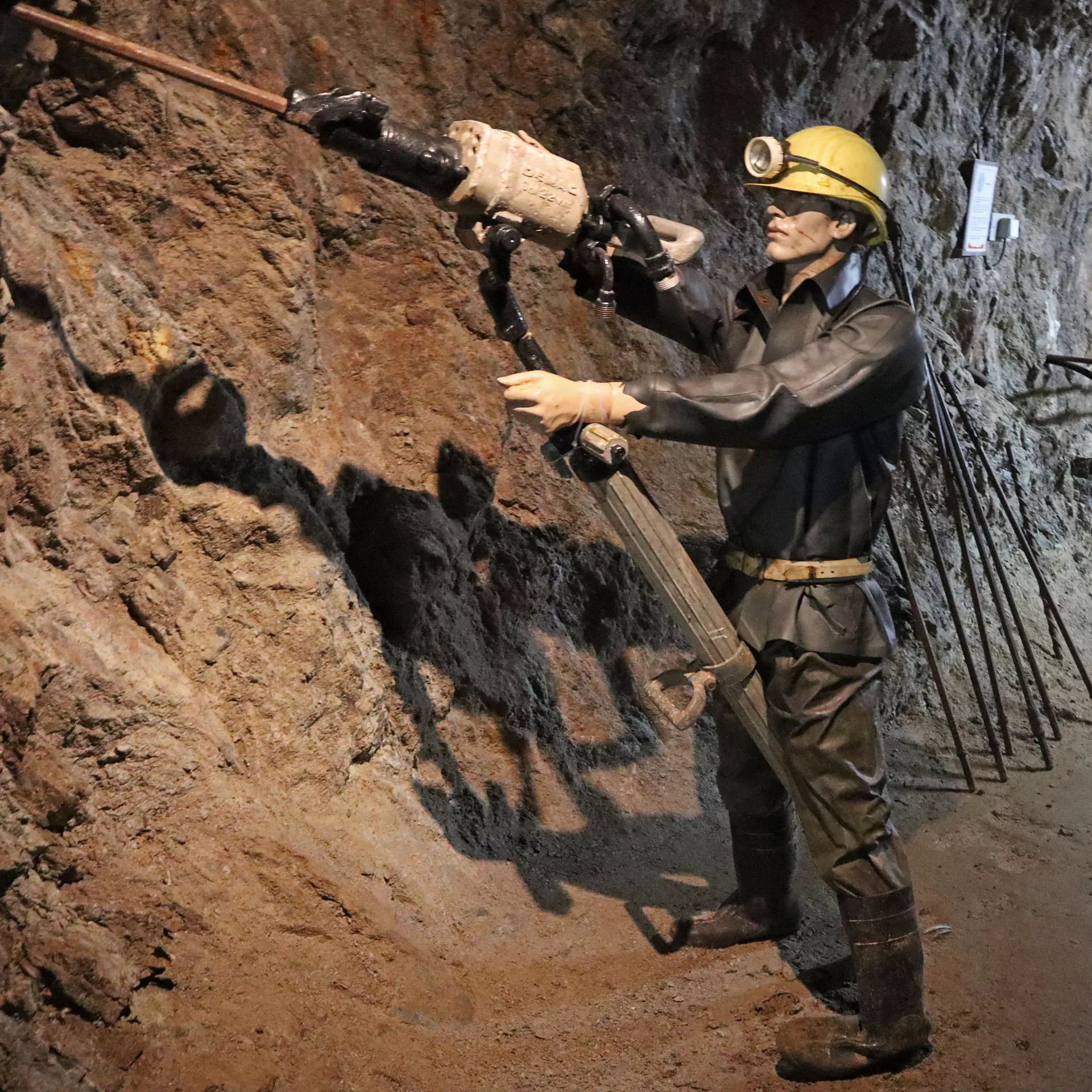 In einem Besucherbergwerk steht als Puppe ein nachgebildeter Bergarbeiter im dunklen Schutzanzug an der Felswand. Er trägt einen gelbem Helm mit aufgesetzter Lampe. In den Händen hält er eine große lange Maschine zum Bohren von Sprengklöchern.