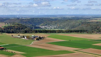 Von erhöhtem Standpunkt aus geht der Blick auf schachbrettartig angelegte Acker- und Grünlandflächen, ehe sich weiter hinten bewaldete Hänge mit bwirtschafteten Ebenen abwechseln. Noch weiter entfernt sind verschiedene Höhenzüge erkennbar.