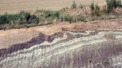 Die Aufschlusswand zeigt unten eine Wechselfolge von rotviolettem und grauweißem Gestein, das oben girlandenartig verformt ist. Darüber liegt ein geringmächtiges hellbraunes Sediment. Hinter einem schmalen Grünstreifen folgt eine Ackerfläche.