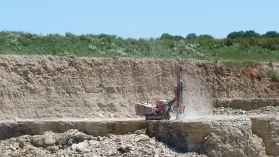 Zu sehen ist ein aktiver Steinbruch in dem ein Bohrgerät, das einem Bagger ähnelt, vertikal eine Bohrung abteuft. 