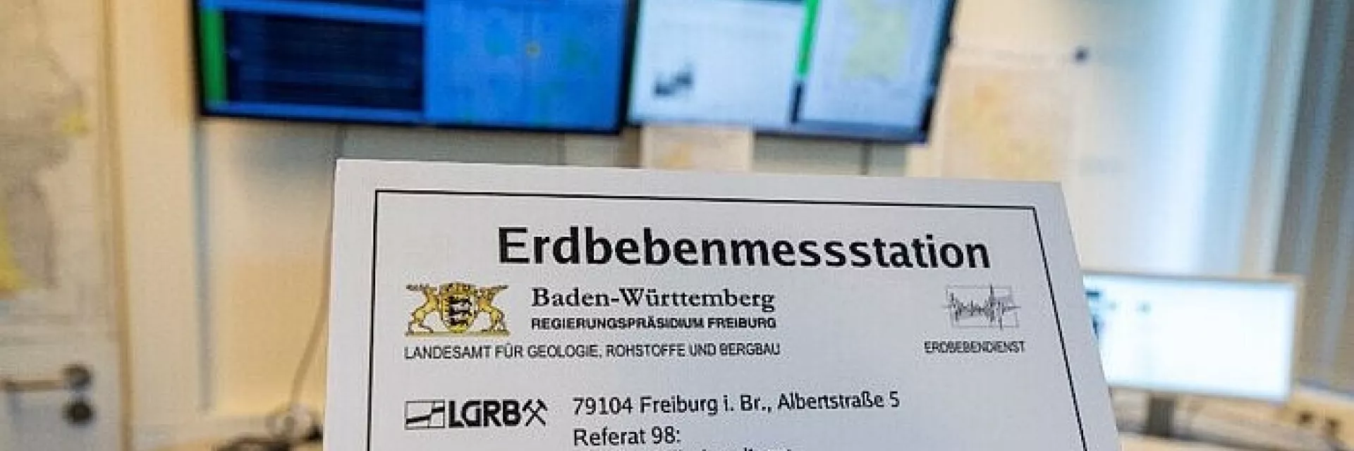 Schild "Erdbebenmessstation" in einem Raum, im Hintergrund mehrere Monitore
