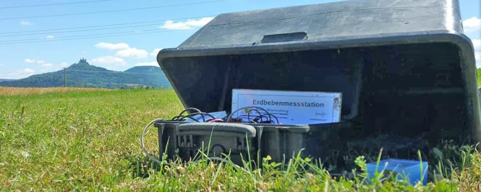 Im Hintergrund links die Burg Hohenzollern. Im Vordergrund steht auf einer Wiese eine temporäre Erdbebenmessstation mit einem blauen Seismometer rechts und einer schwarzen Box mit Kabeln in der Mitte, beides wird von einer umgedrehten schwarzen Kunststoffwanne geschützt.