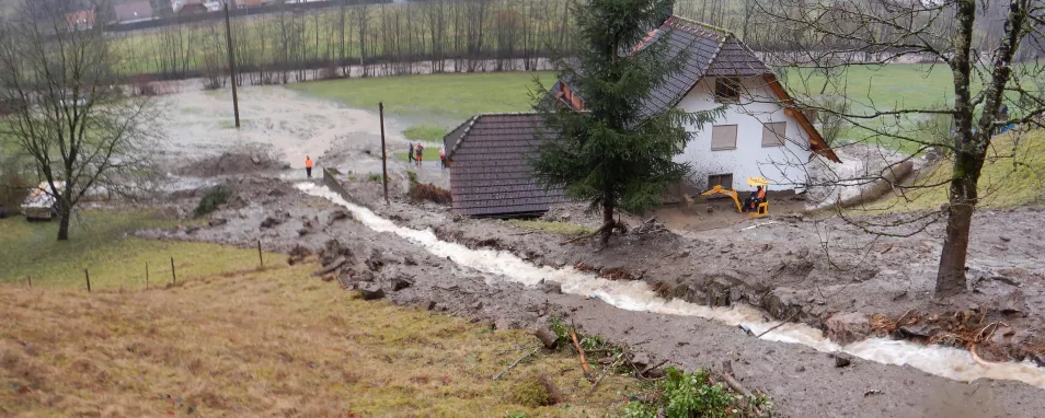 Blick hangabwärts mit Ansicht eines kleinen, rauschenden Bachs. An beiden Uferseiten sind Schlamm, Steine und Baumstämme angehäuft. Angrenzend zum Bach befindet sich ein mit Schlamm verschmutztes Wohngebäude. 