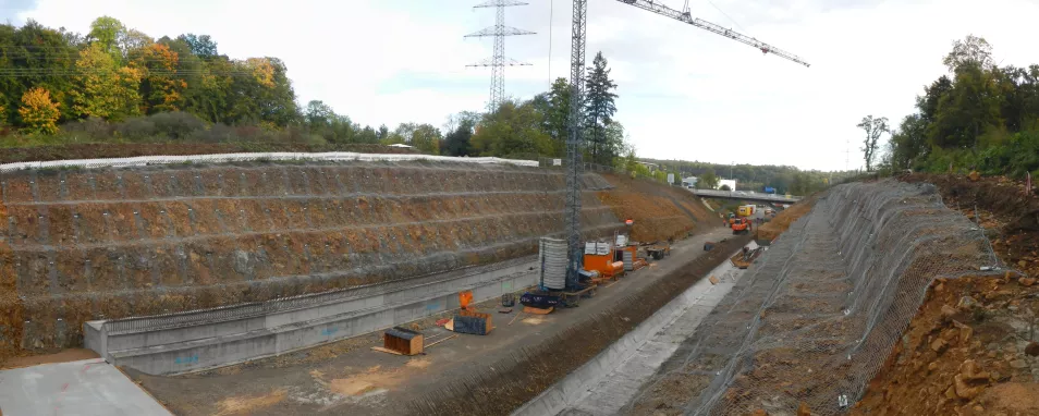 Blick auf eine Baustelle an einem länglichen Einschnitt in einem Hanggelände. In der Bildmitte sind eine im Bau befindliche Straße sowie ein Kran und weitere Baustelleneinrichtungen zu erkennen. Zu beiden Seiten der Straße ist die Hang künstlich abgetreppt. An den Stufen sind ockerfarbener bis brauner Boden und Steine zu sehen. Diese sind mit grobmaschigen Netzen abgedeckt.