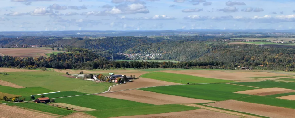 Von erhöhtem Standpunkt aus geht der Blick auf schachbrettartig angelegte Acker- und Grünlandflächen, ehe sich weiter hinten bewaldete Hänge mit bwirtschafteten Ebenen abwechseln. Noch weiter entfernt sind verschiedene Höhenzüge erkennbar.