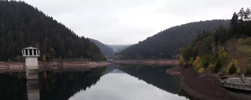 Blick auf eine dunkle Wasserfläche im Vordergrund und bewaldete Hügel im Hintergrund. Der wolkenverhangene graue Himmel spiegelt sich in der Wasseroberfläche. Technische Anlagen eines Stausees sind an beiden Bildrändern zu erkennen. Unbewachsene Uferböschungen zeigen den maximalen Aufstau des Sees an. 