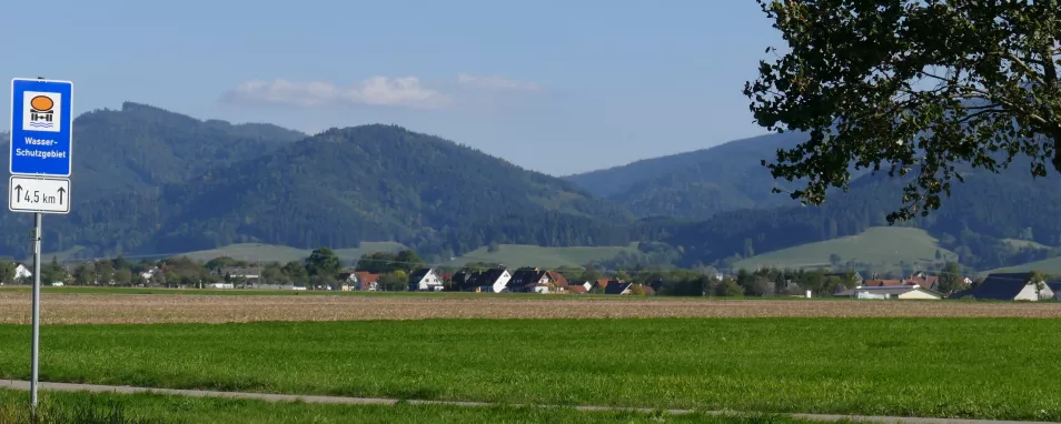 Schild mit Aufschrift "Wasserschutzgebiet". Im Hintergrund Wiesen- und Ackerflächen sowie bewaldete Hänge des Schwarzwalds.