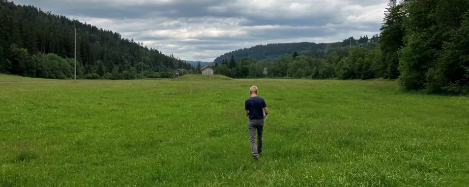 Foto zeigt einen Mann, der über eine mit Bäumen gesäumte Wiese auf ein Brunnenhaus zugeht